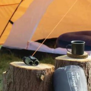 A camping scene with an orange tent in the background. In the foreground, two tree stumps serve as stools or tables, holding various camping accessories like a mug, binoculars, and a coiled red rope. A sleeping bag in a sack is also next to the stumps.