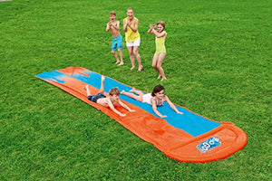 A group of kids playing on a water slide at an outdoor playground, with some lying on blue and orange mats.